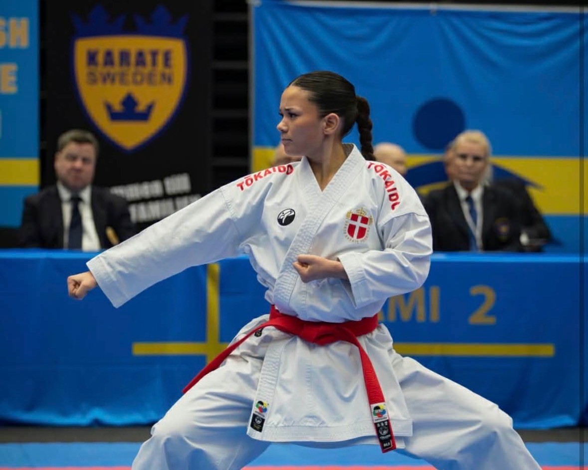 Freja Chung Jepsen in a deep kata stance with a red belt in front of the judges' table at a karate tournament