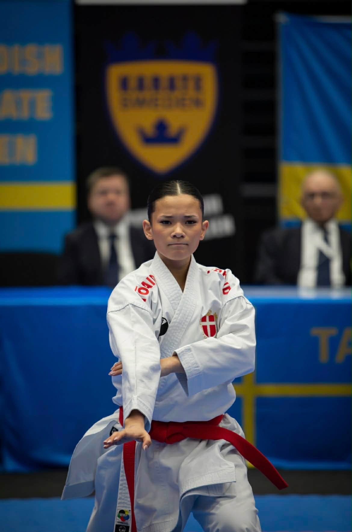 Freja Chung Jepsen focused in a kata stance, wearing a red belt and the Danish crest, in front of the judges at a karate championship