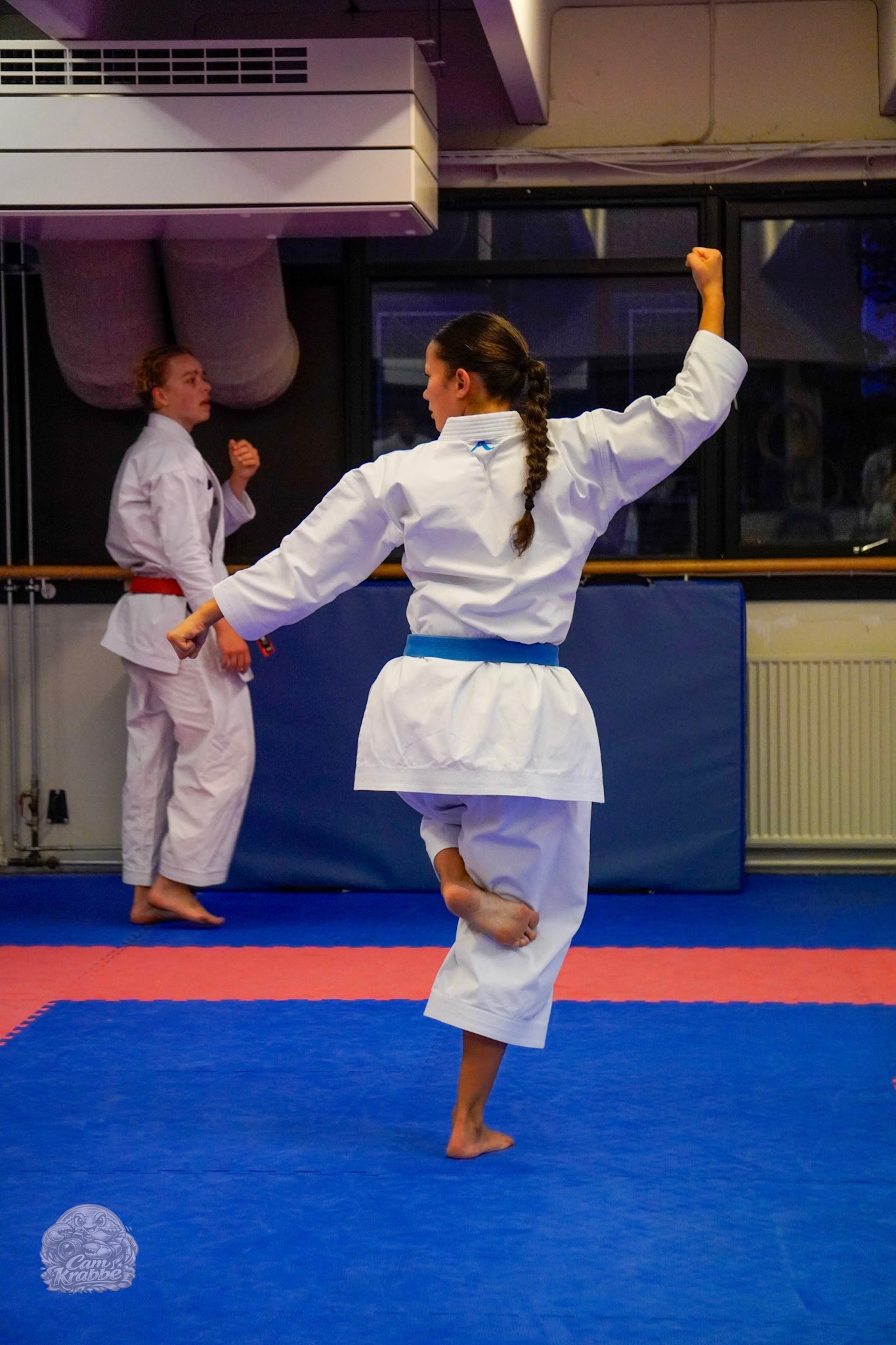 Freja Chung Jepsen balancing on one leg with a raised fist during a kata at the training hall, seen from behind