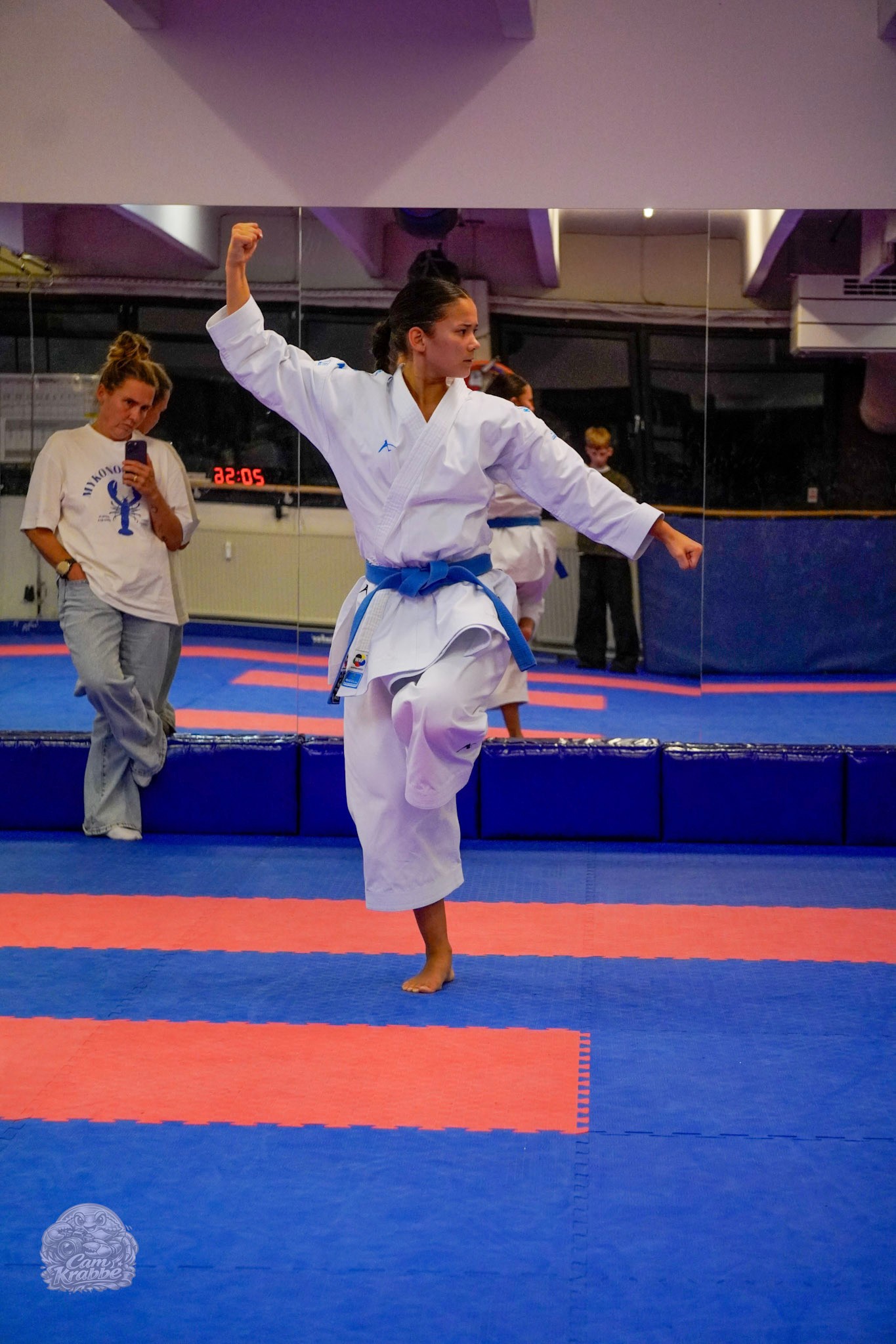 Freja Chung Jepsen balancing on one leg in a blue belt during kata training in front of the dojo mirrors